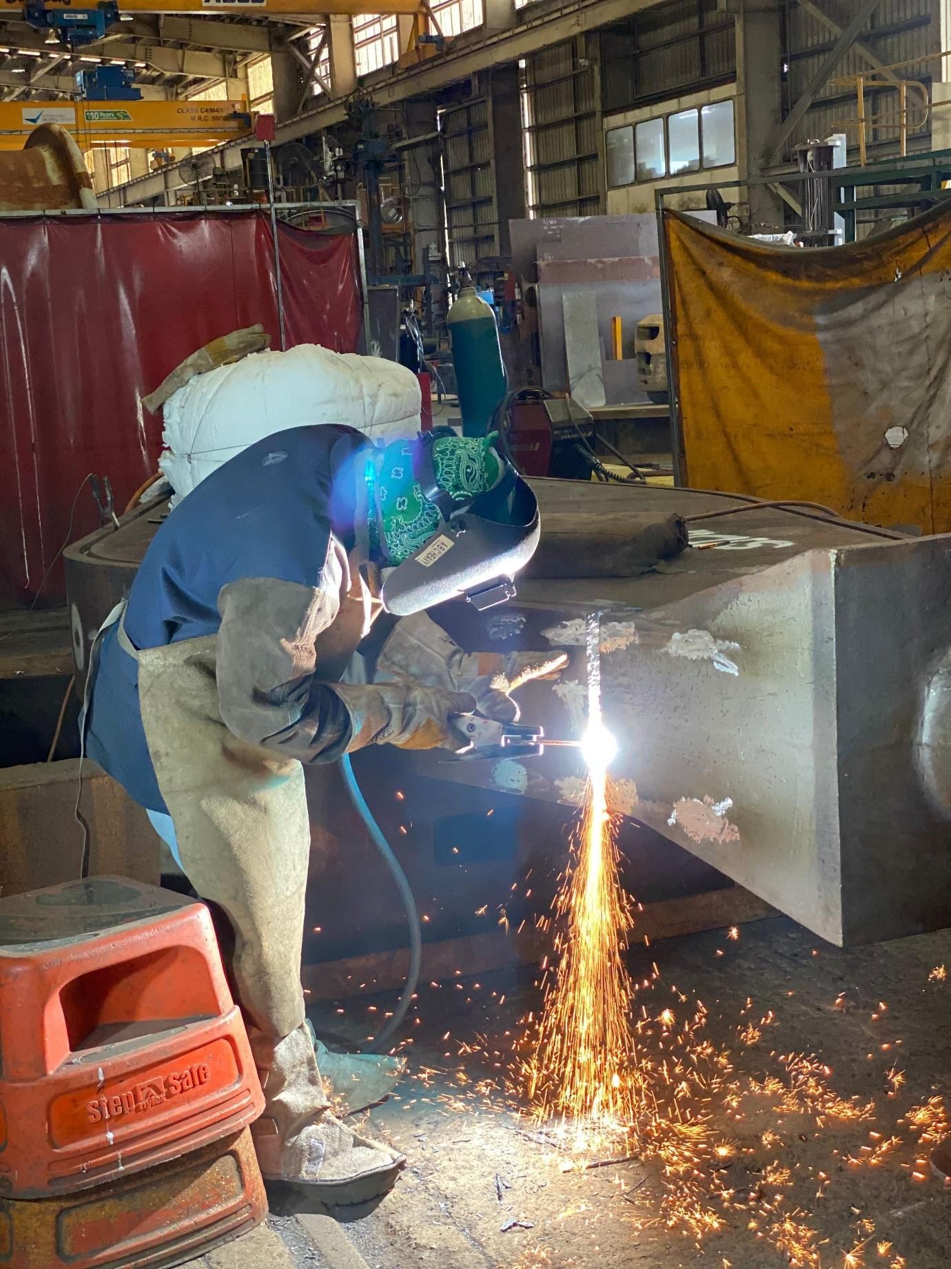 ABC welder in full mask cutting a heavy fabricated steel component, blue-green arc light and sparks in the fabrication shop