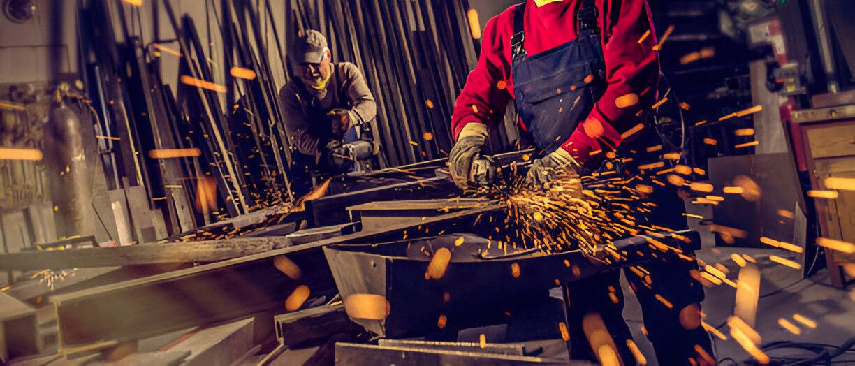 Two ABC boilermakers dressing a heavy steel weldment, grinding sparks flying across the fabrication bay