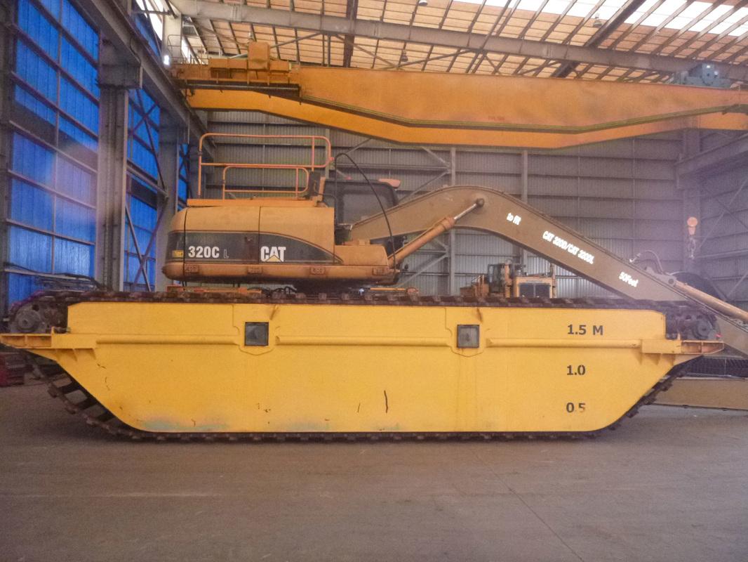 A CAT 320C excavator mounted on a custom amphibious pontoon undercarriage inside the ABC workshop, beneath the overhead gantry crane
