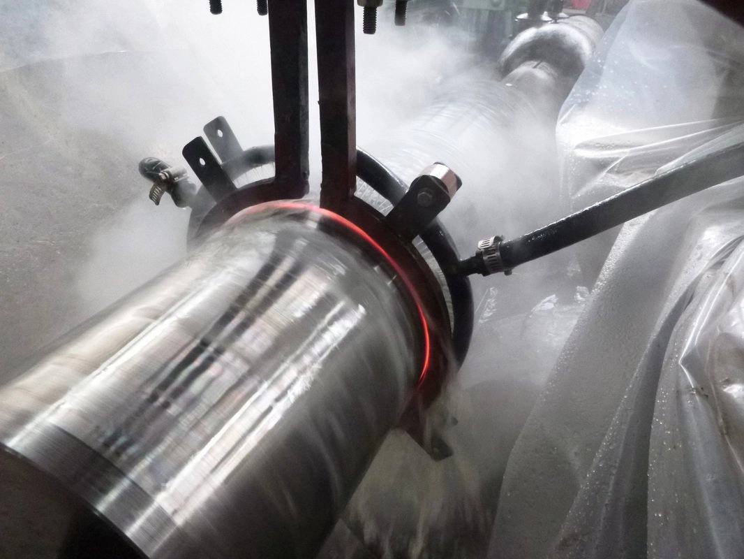 Induction coil glowing red around a rotating steel shaft as quench mist rises in the heat-treatment bay