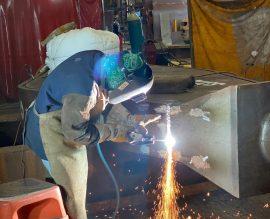 ABC welder in protective gear mid-arc on a fabricated steel section, sparks falling to the floor