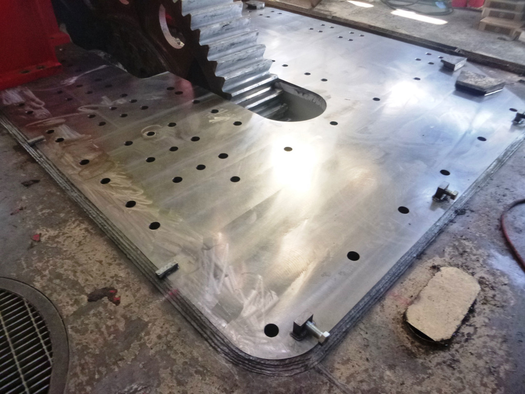 Freshly machined dragline baseplate catching the light on the workshop floor, a large gear resting at its edge